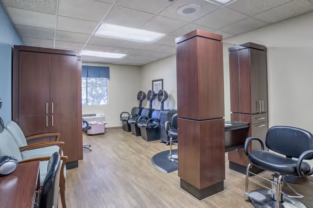 Interior view of a salon area in a senior living facility with wooden cabinets, salon chairs, hair dryers, and a window with blinds letting in natural light.