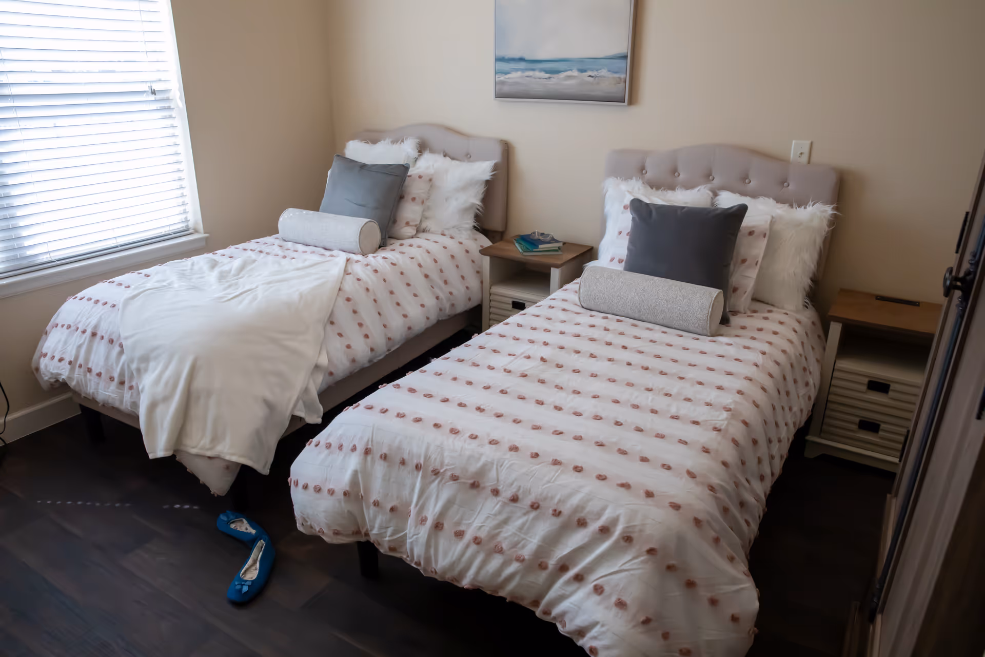 A cozy bedroom with two twin beds featuring tufted beige headboards and white bedding with pink polka dots. Each bed has a gray pillow, a cylindrical bolster pillow, and fluffy white accent pillows. A small wooden nightstand with books and glasses sits between the beds. A window with white blinds is on the left wall, and a painting of a seascape hangs above the beds. A pair of blue slippers is on the dark wooden floor near the left bed.