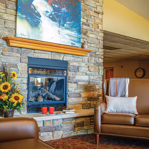Cozy assisted-living common area with a stone fireplace flanked by leather armchairs, throw pillows, artwork, and a vase of sunflowers.