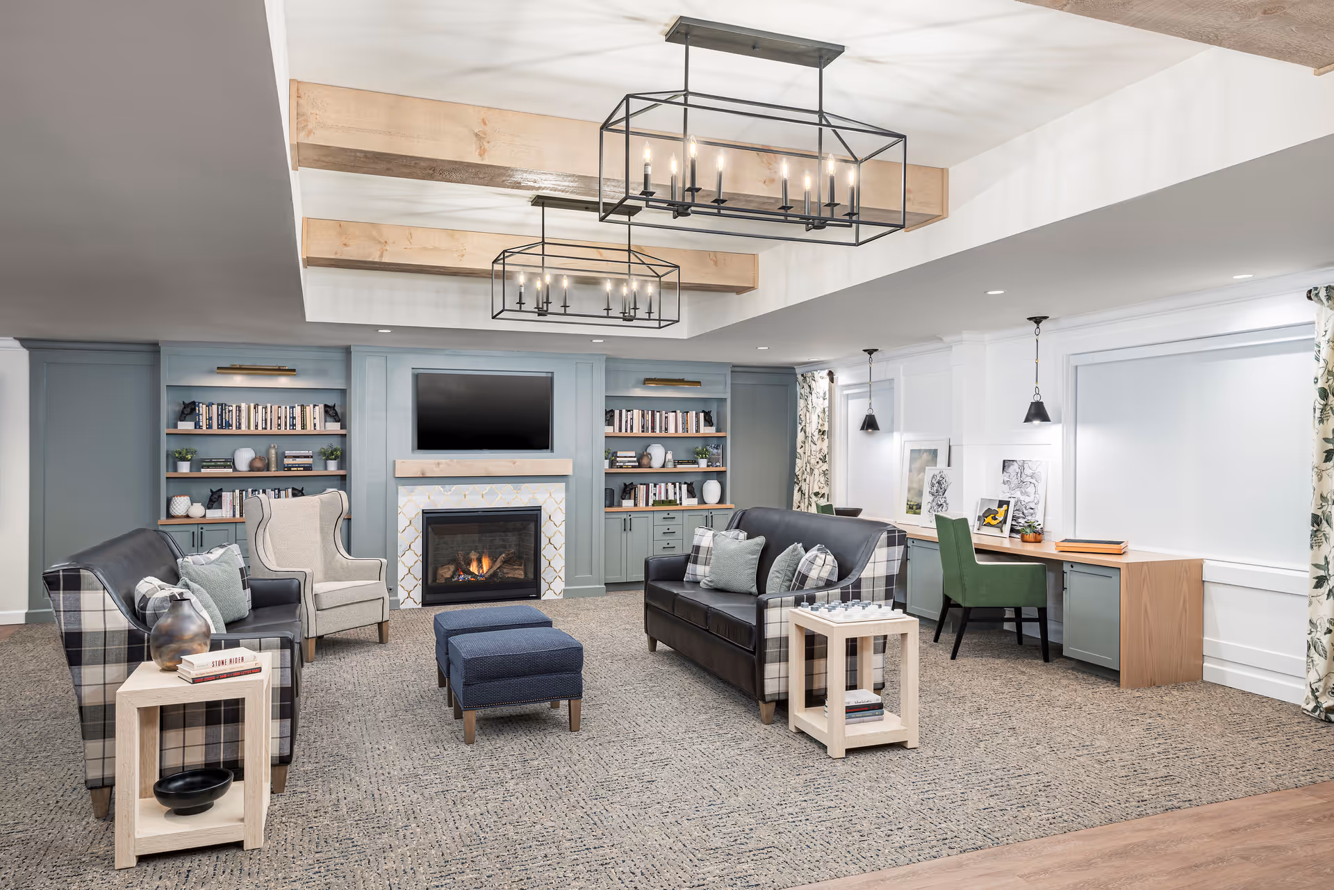 A cozy living room area with two plaid armchairs, a gray wingback chair, and a dark leather sofa with plaid accents. The room features a fireplace with a TV mounted above it, built-in shelves filled with books and decorative items, and two modern rectangular chandeliers hanging from a ceiling with exposed wooden beams. There is a long desk with green chairs and framed artwork on the right side, and the floor is carpeted with a neutral tone.