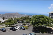 Parking lot with cars, green trees, and a distant coastal hillside under a blue sky.