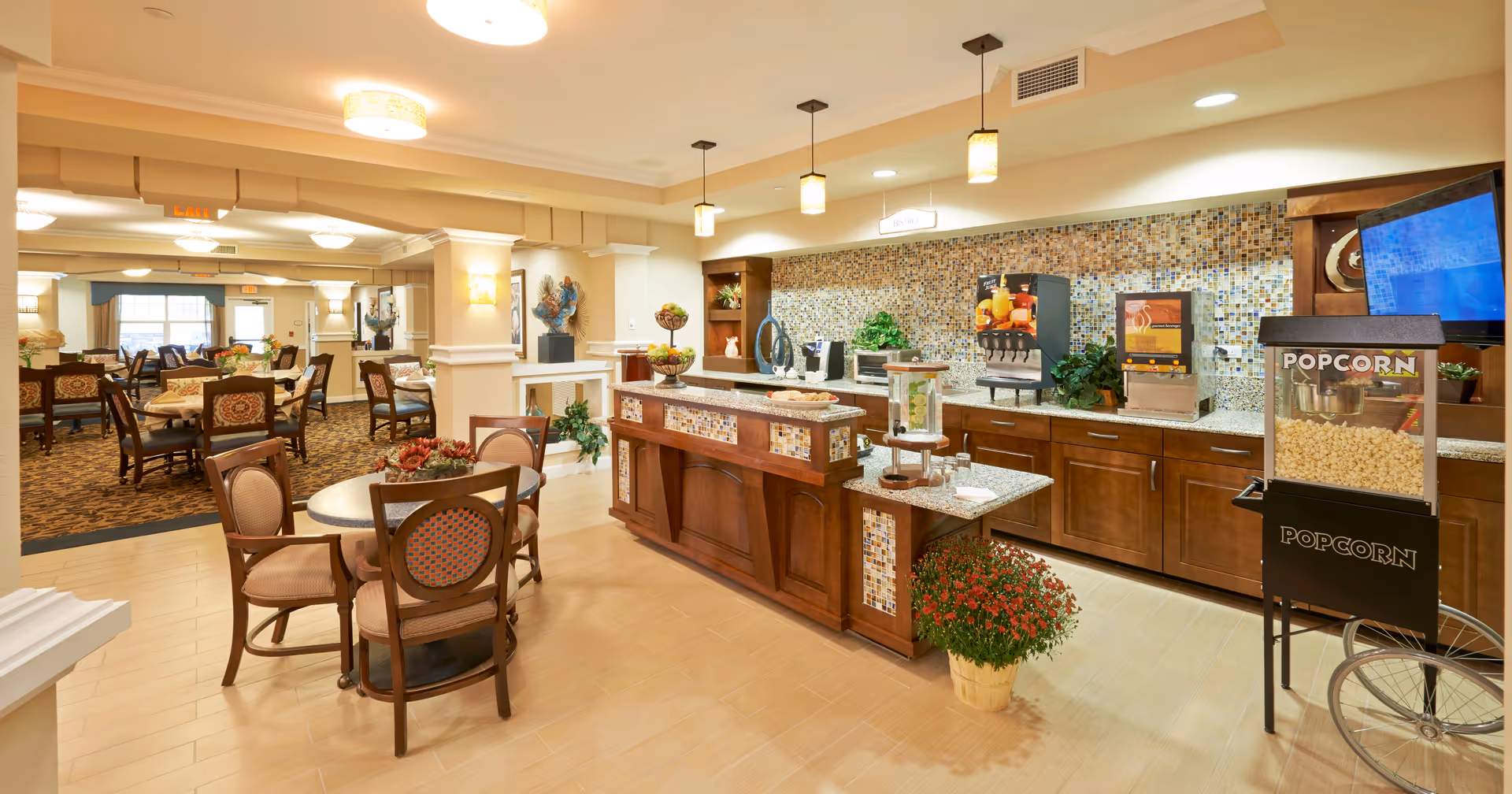 A bright and spacious dining area in an assisted living facility featuring multiple round tables with chairs, a central island with snacks and beverages, a popcorn machine, and a beverage dispenser against a mosaic tiled wall. The room has warm lighting and a carpeted area with additional seating in the background.