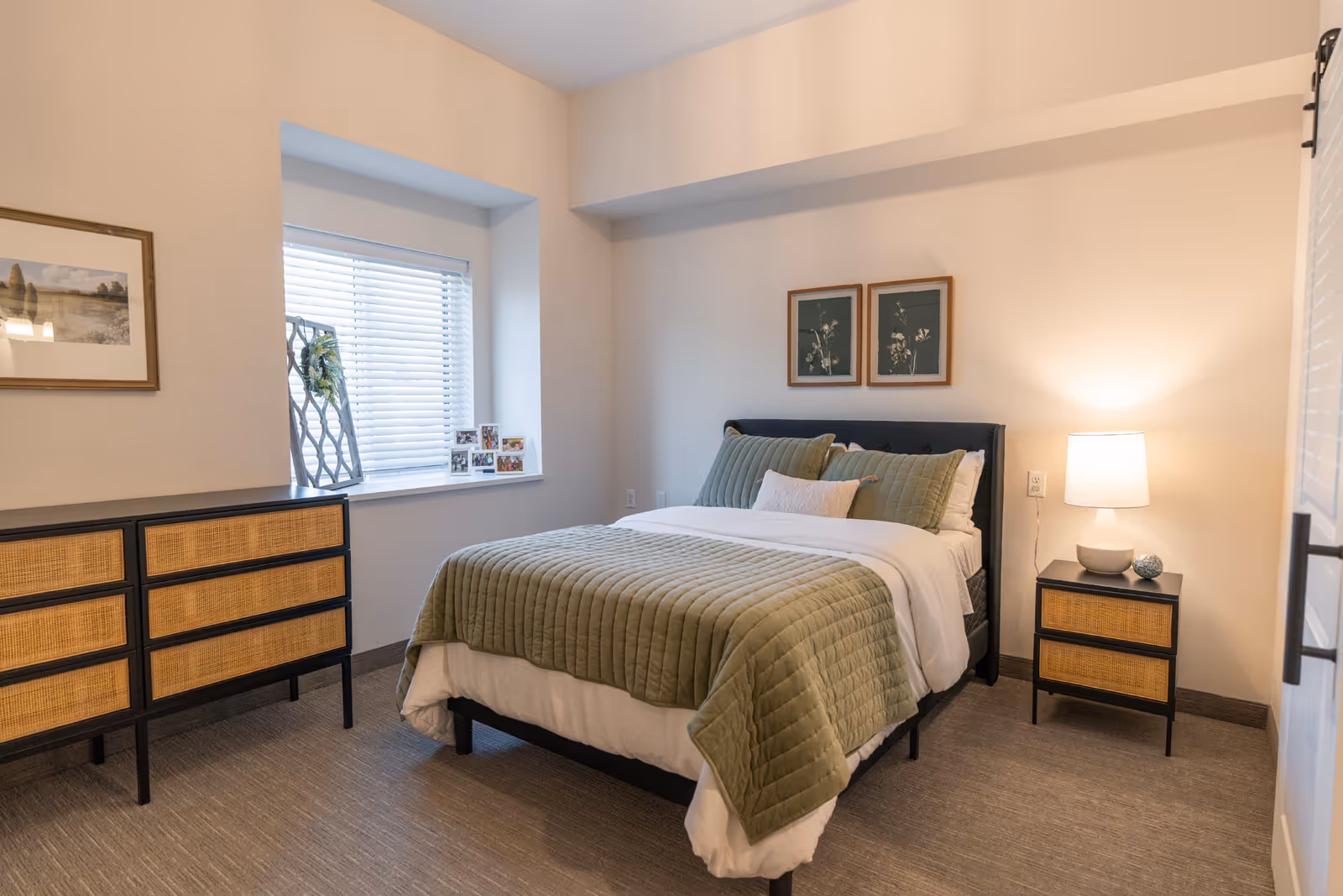 A neatly staged bedroom with a bed dressed in a green quilt and pillows, a nightstand with a lamp, and a dresser beneath a window.