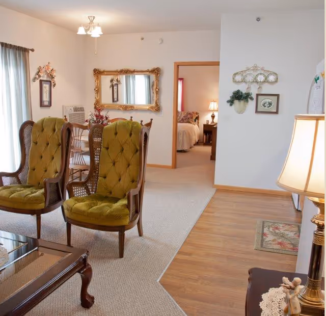Interior view of a senior living facility showing a living room with two green upholstered armchairs, a wooden coffee table, and a dining area with a table and chairs in the background. A hallway leads to a bedroom with a bed and a lit bedside lamp. The walls are decorated with framed pictures and a large ornate mirror.