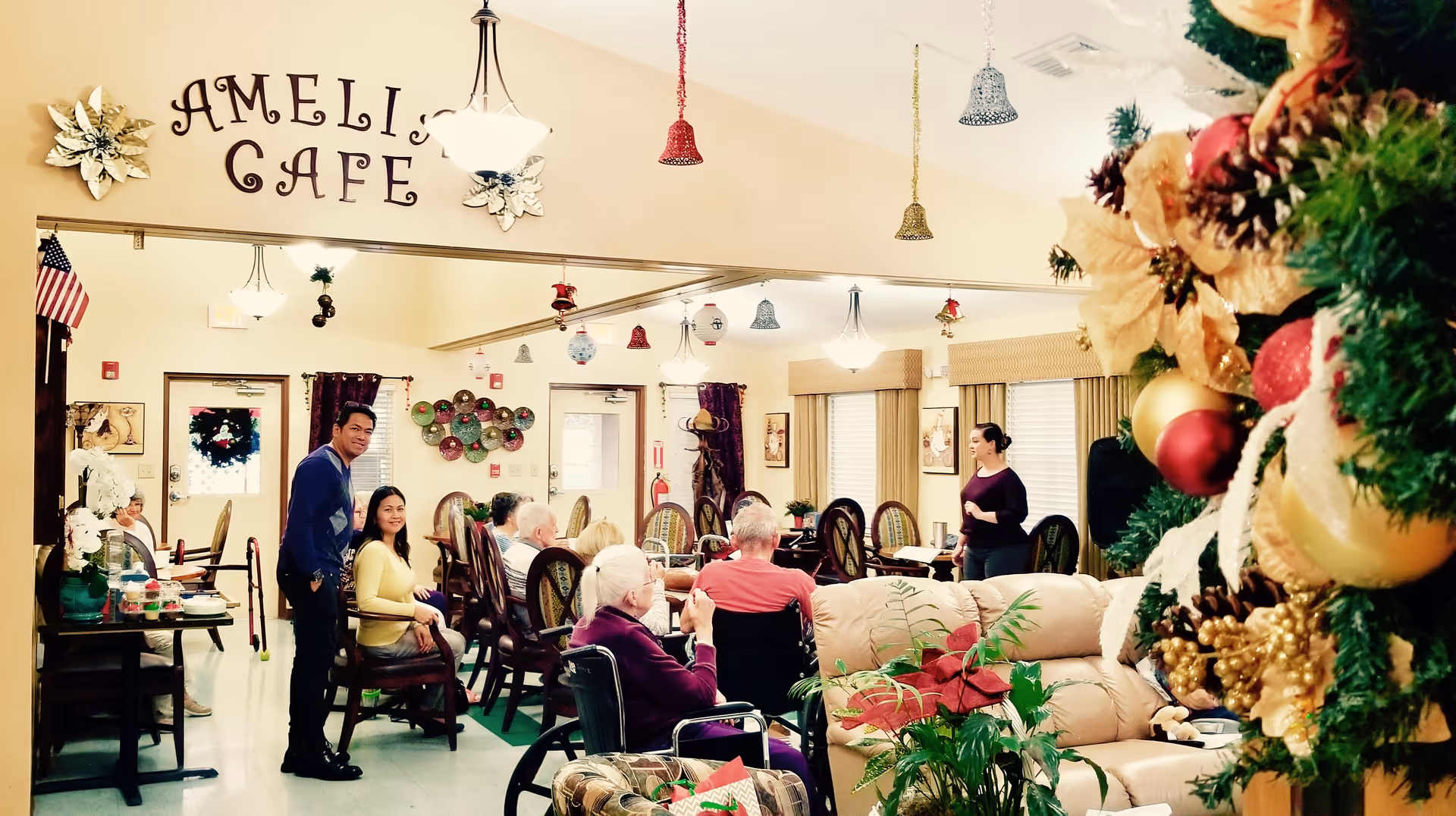 Residents and staff gathered in a decorated communal dining/lounge area labeled "Ameli's Cafe" with holiday decorations.
