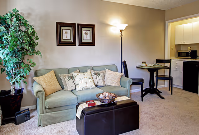 A cozy living room area with a light green sofa adorned with palm tree patterned and solid color pillows. There is a tall floor lamp beside the sofa and two framed palm tree artworks on the beige wall above it. A dark brown ottoman with a decorative bowl and a small red box sits in front of the sofa. To the right, a small round dining table with two chairs is set near a kitchenette with white cabinets, a microwave, and a mini fridge. A large potted plant is placed on the left side of the sofa.