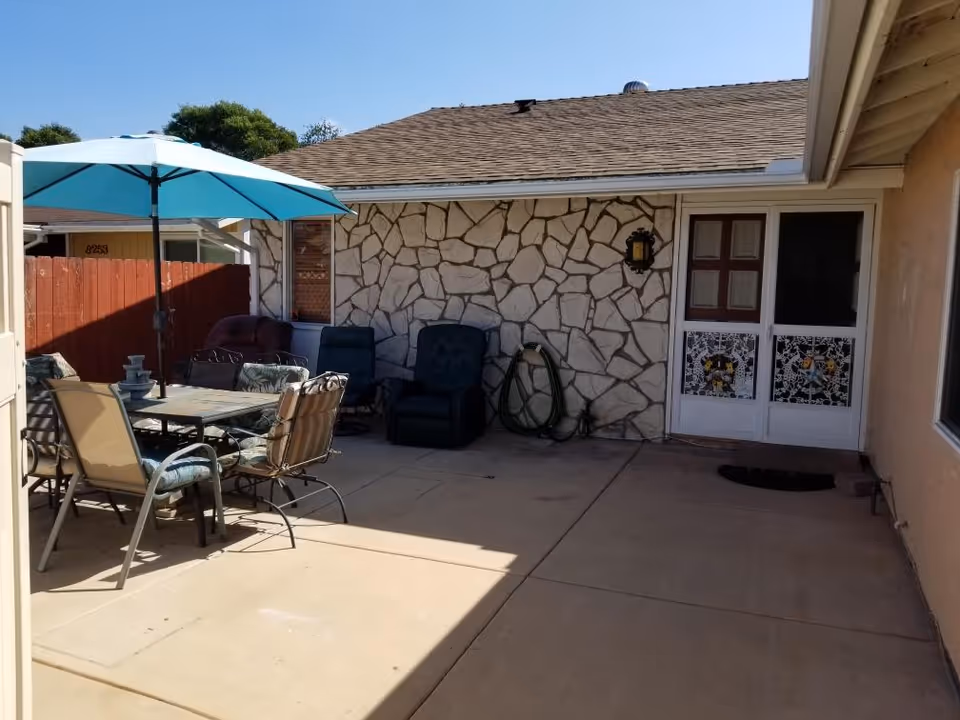 Sunlit fenced backyard patio with a dining table and umbrella, lounge chairs, a coiled hose, and a stone-faced house wall with a screen door.
