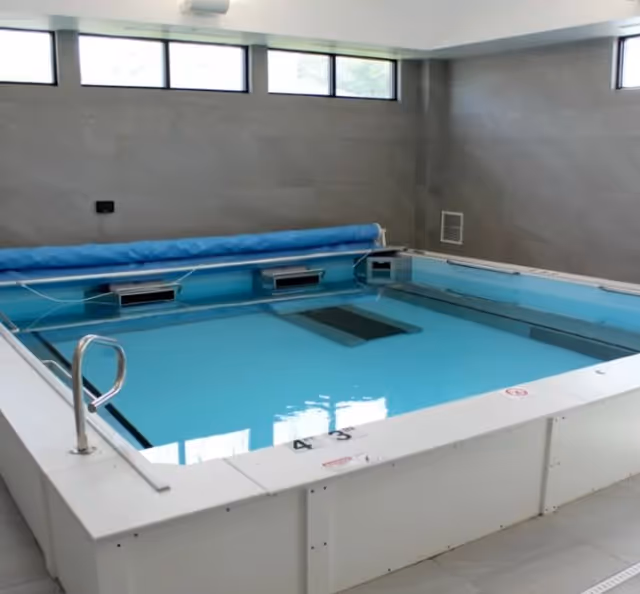 Indoor therapy pool with clear blue water, surrounded by white tiled walls and floor, with a metal handrail on the left side and windows near the ceiling letting in natural light.