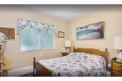 A cozy bedroom with a wooden bed frame and a floral bedspread in shades of purple and white. The room has beige walls, a window with floral curtains, a nightstand with a lamp, and a framed picture hanging above the bed.