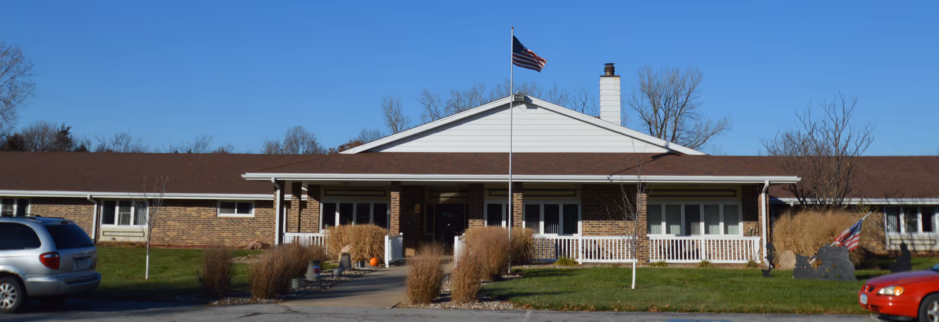 Front exterior view of Ridgewood Specialty Care building with a flagpole displaying an American flag in the center. The building has a brick facade with white siding on the upper portion and a brown roof. There are some bushes and grass in front, along with a sidewalk leading to the entrance. Two cars are partially visible on either side of the image.