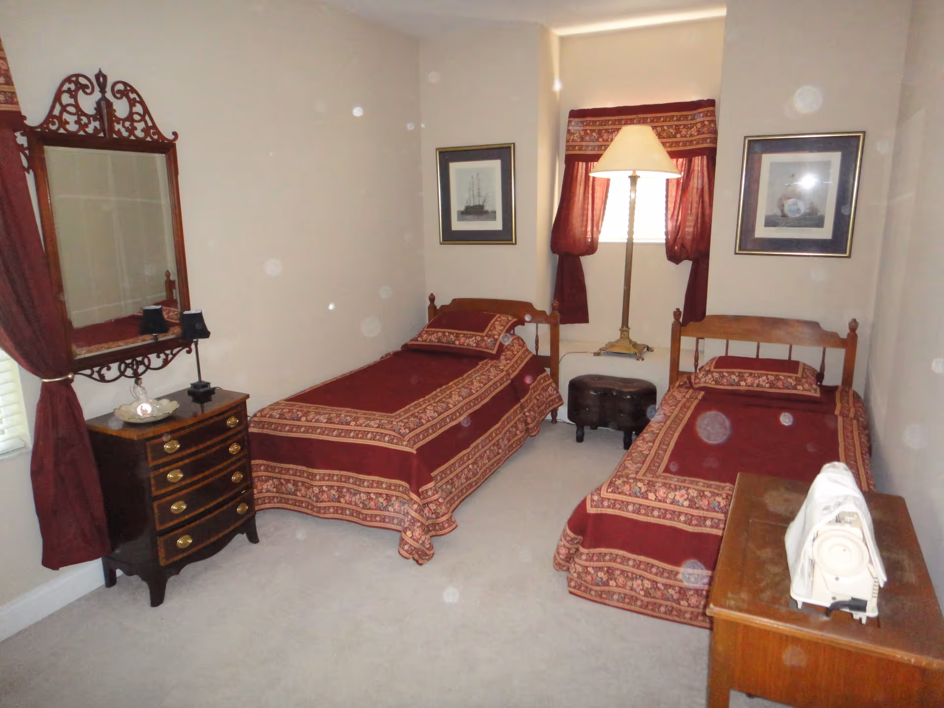 A bedroom with two single beds covered in matching red and gold patterned bedspreads. Between the beds is a small round ottoman and a tall floor lamp. On the left side of the room is a wooden dresser with a decorative mirror above it and a small lamp. The window has red curtains that match the bedspreads. There are two framed pictures on the wall above the beds.