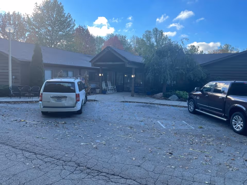 Exterior view of Chalet Rehabilitation and Healthcare Center showing the entrance with a covered porch, two parked vehicles in front, some outdoor seating to the left, and trees in the background under a partly cloudy sky.