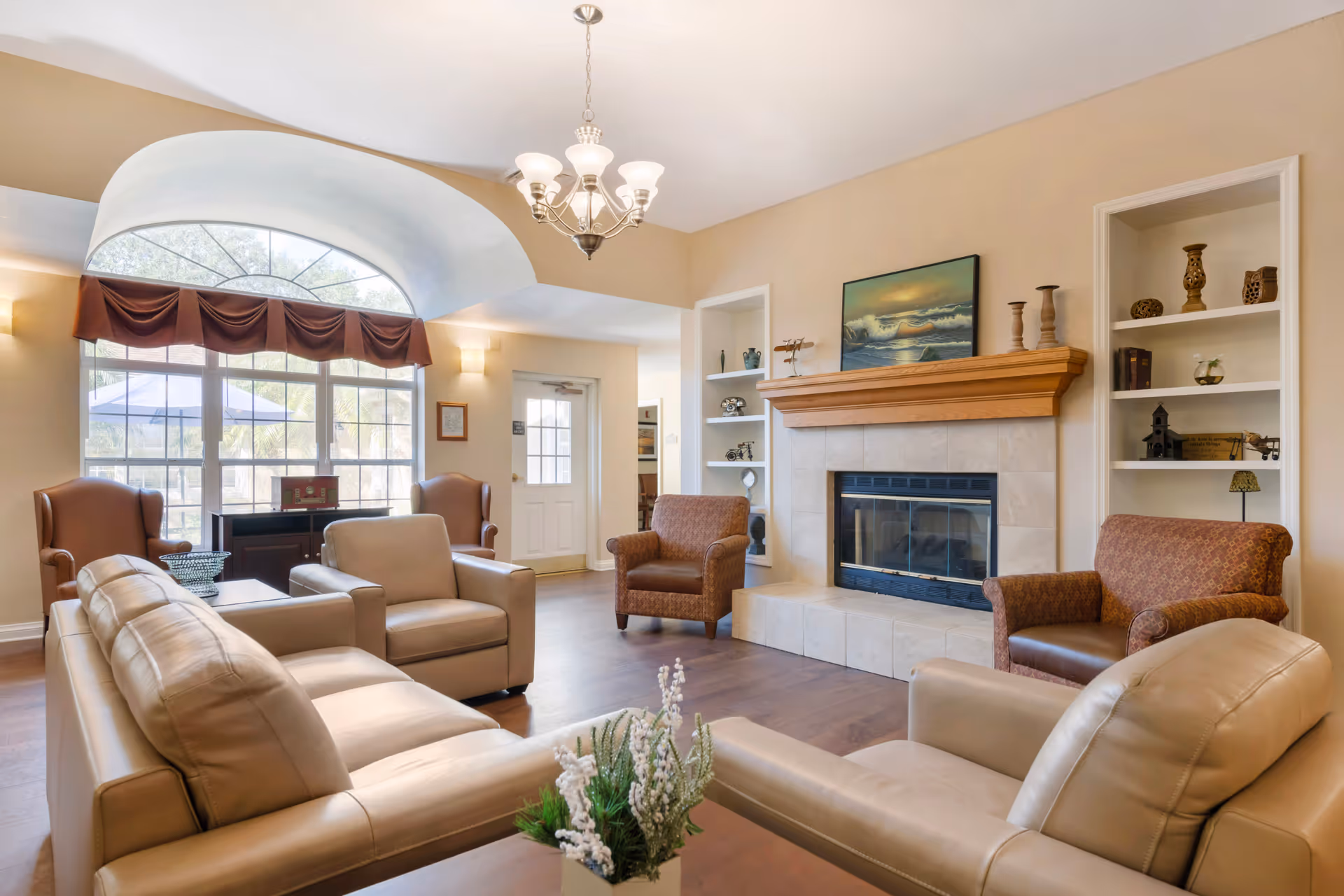 A cozy living room area with beige leather sofas and armchairs arranged around a fireplace with a wooden mantle. The room features built-in shelves with decorative items, a large arched window with a brown valance, and a chandelier hanging from the ceiling. The walls are painted a warm beige color and the floor is wooden.