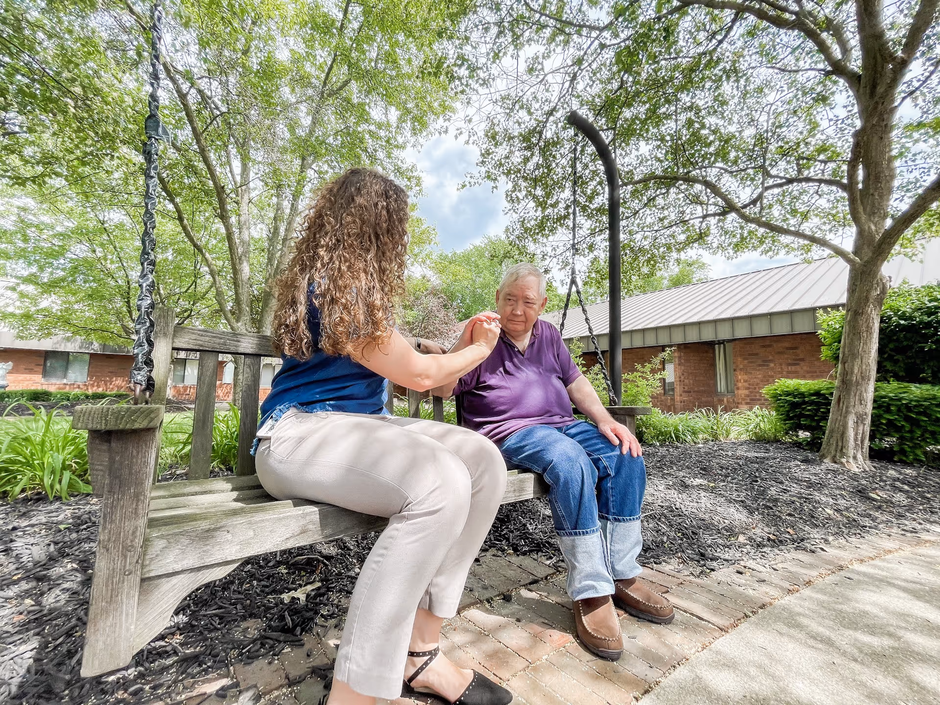 An elderly man and a woman sitting on a wooden swing bench in a garden area outside a brick building. The woman is holding the man's hand and they appear to be engaged in a caring conversation. Trees and greenery surround them under a partly cloudy sky.