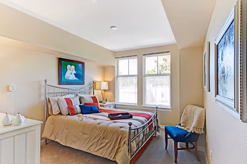 A cozy bedroom with a metal frame bed dressed in beige and red bedding, multiple pillows, and a tray on top. There is a nightstand with a lamp and clock beside the bed, a blue cushioned chair with a beige throw blanket, and two large windows letting in natural light. Artwork hangs above the bed and on the adjacent wall.