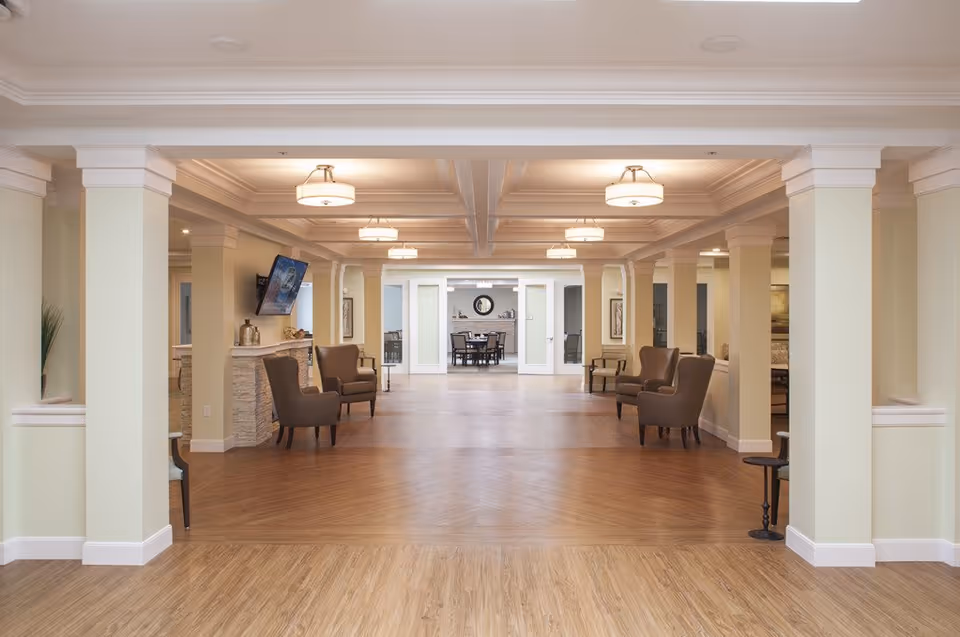Spacious senior living common lounge with columns, armchairs, overhead lights, and a dining area visible in the background.