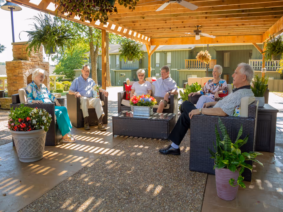 A group of six elderly people sitting and chatting under a wooden pergola in an outdoor patio area. They are seated on wicker chairs around a glass-top coffee table with a flower arrangement. The setting includes hanging plants, potted plants, and a stone fireplace in the background, with a residential building visible behind them.