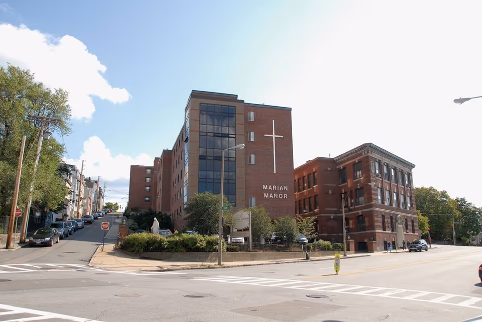 Exterior view of Marian Manor Nursing Home, a multi-story brick building with large windows and a prominent white cross on the front. The building is situated at a street corner with a few cars parked along the road and some greenery in front.