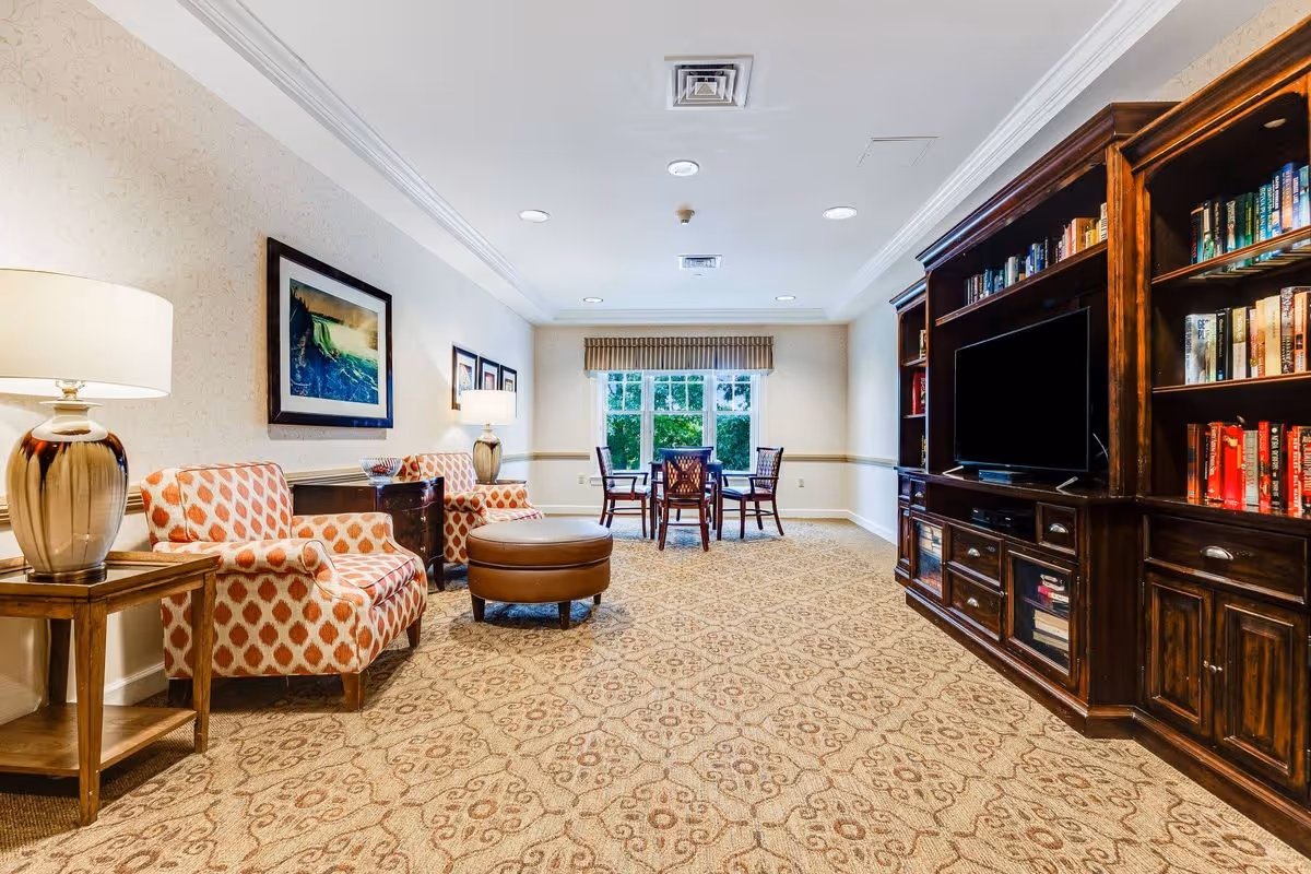 Bright common living area with patterned armchairs, an ottoman, lamps, a large wooden entertainment bookshelf with a TV, and a dining table by the window.