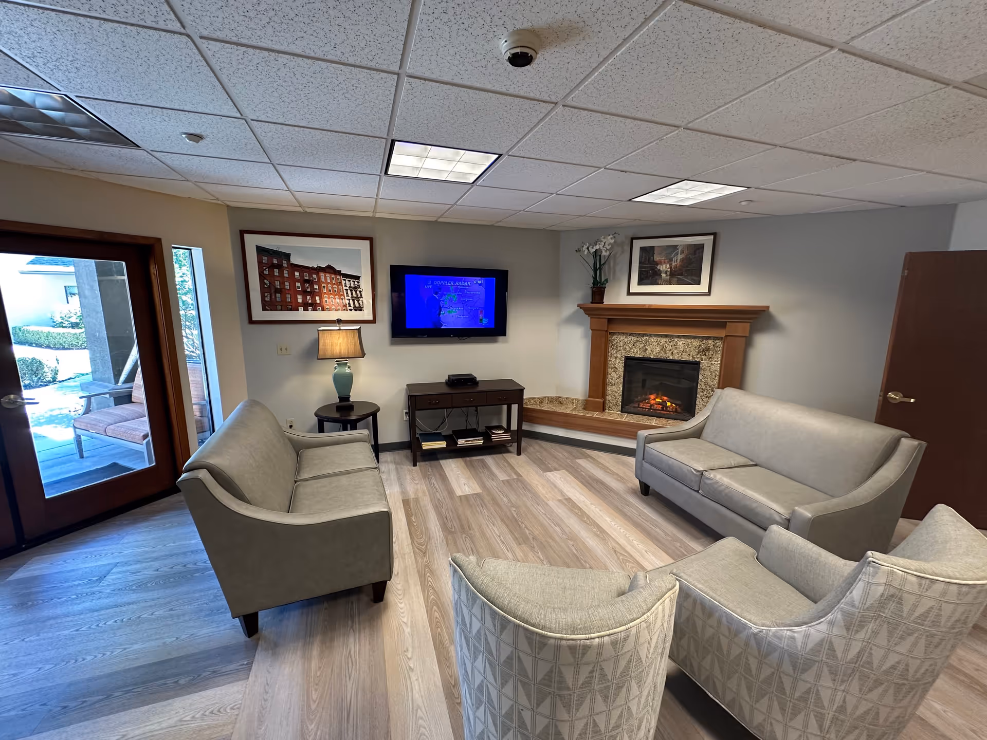A cozy living room area with two gray sofas, two patterned armchairs, a wooden side table with a lamp, a wall-mounted TV, and a fireplace with a wooden mantel. There are framed pictures on the walls and a glass door leading to an outdoor patio with seating.