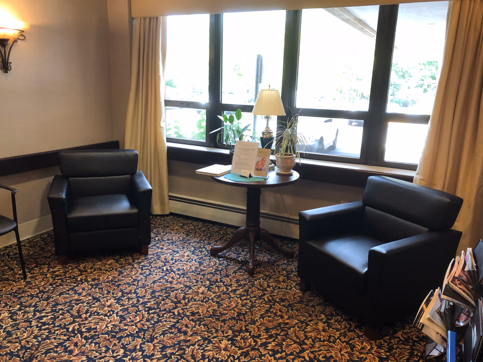 A cozy sitting area with two black armchairs positioned around a small round wooden table. The table holds a lamp, two potted plants, and some papers. The room has a large window with beige curtains, and the floor is covered with a patterned carpet featuring autumn leaves.