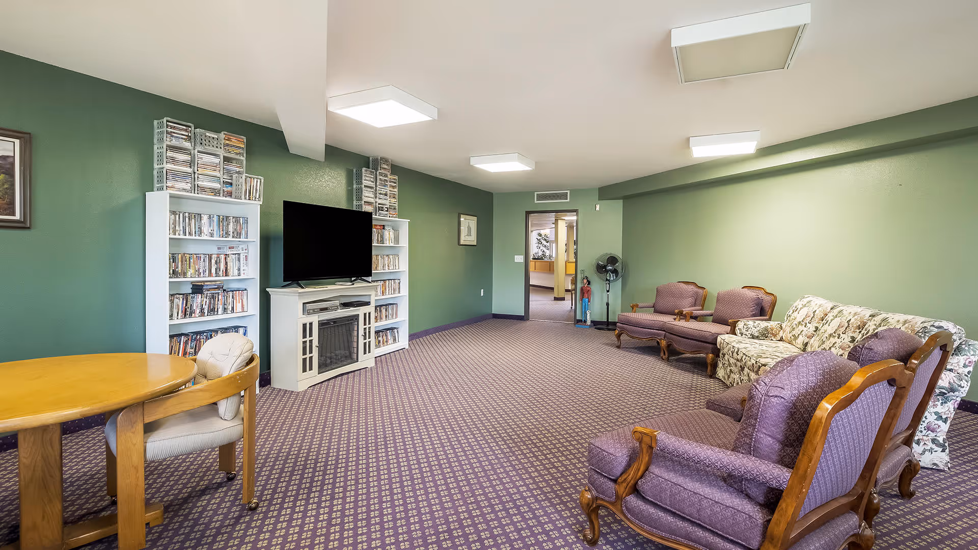 Community lounge with a TV and bookshelves, a round table, and upholstered chairs and sofas on patterned carpet.