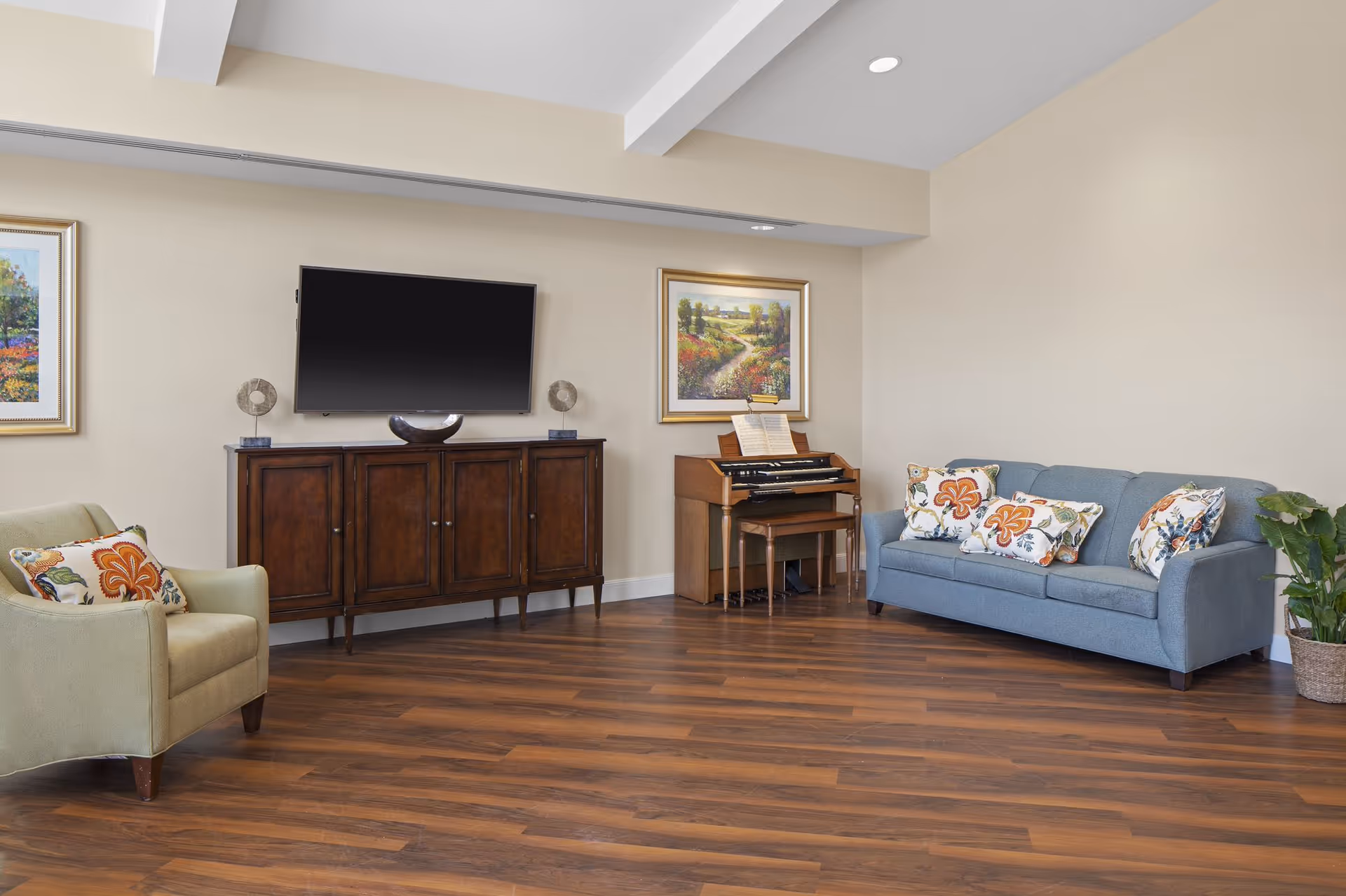 A living room with a blue sofa adorned with floral patterned pillows, a green armchair with a floral pillow, a wooden cabinet with a flat-screen TV mounted above it, and a small wooden organ with a bench. The room has wooden flooring, beige walls, two framed landscape paintings, and a potted plant in the corner.