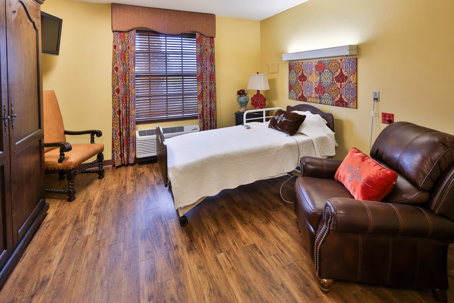 A cozy bedroom in a senior living facility with a single bed covered in a white quilt, a brown leather armchair with a red decorative pillow, a wooden armoire, an upholstered chair, and a window with colorful patterned curtains. The walls are painted yellow and the floor has wood-like flooring.