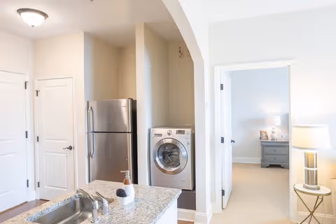 Interior view of a modern kitchen area with a granite countertop island featuring a sink and soap dispenser. In the background, there is a stainless steel refrigerator and a front-loading washing machine. To the right, an open doorway leads to a bedroom with a nightstand, lamp, and framed photos.