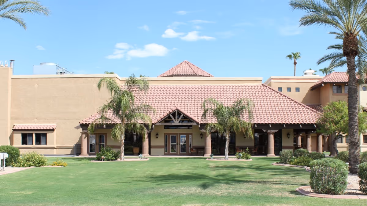 Exterior view of The Forum at Desert Harbor building with a tiled roof, beige walls, palm trees, and a well-maintained green lawn under a blue sky with some clouds.