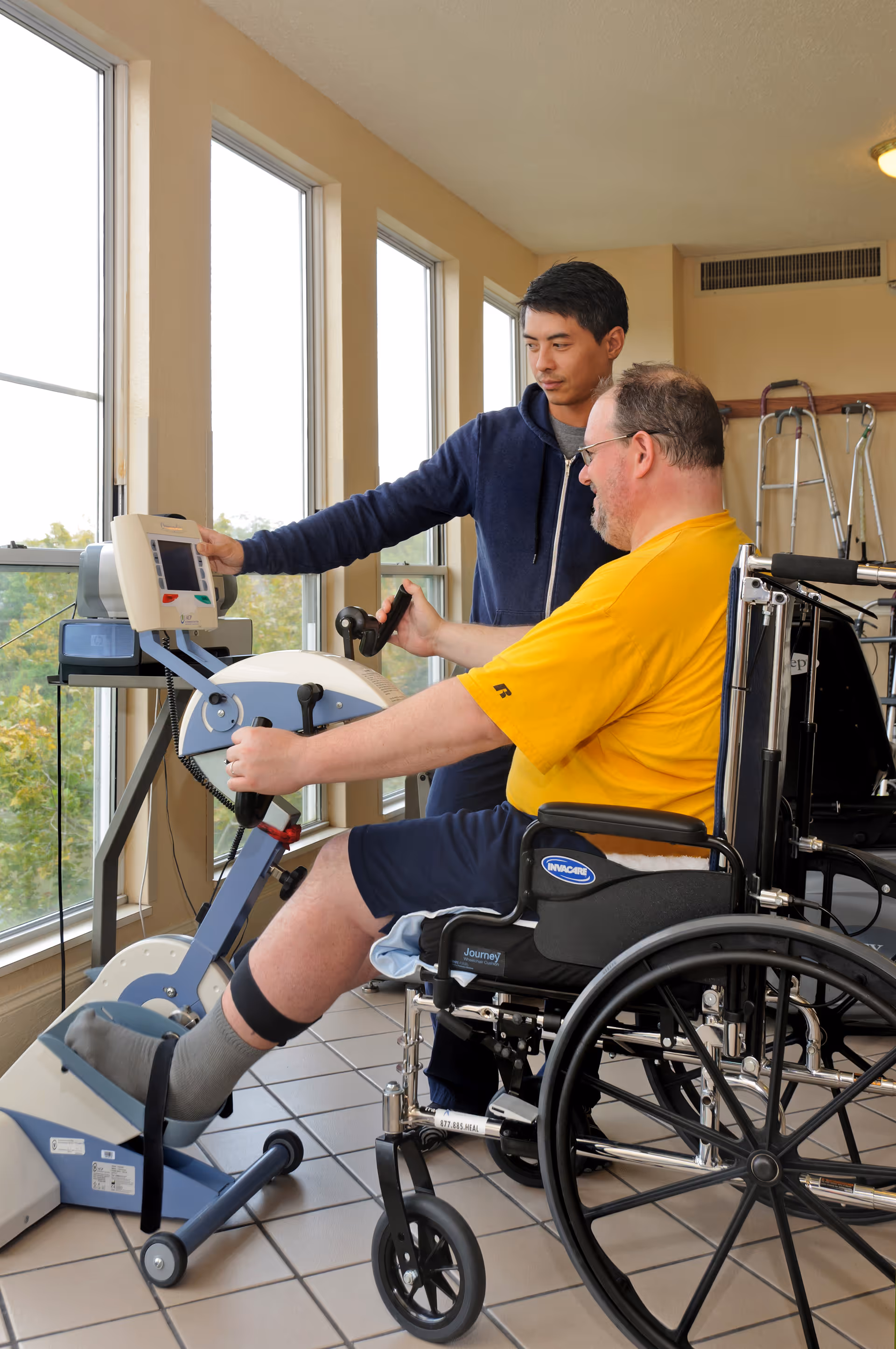 A man in a wheelchair wearing a yellow shirt is exercising his leg on a stationary rehabilitation bike while another man in a blue hoodie assists him by adjusting the machine's settings. They are in a room with large windows and tiled floors, with walkers visible in the background.