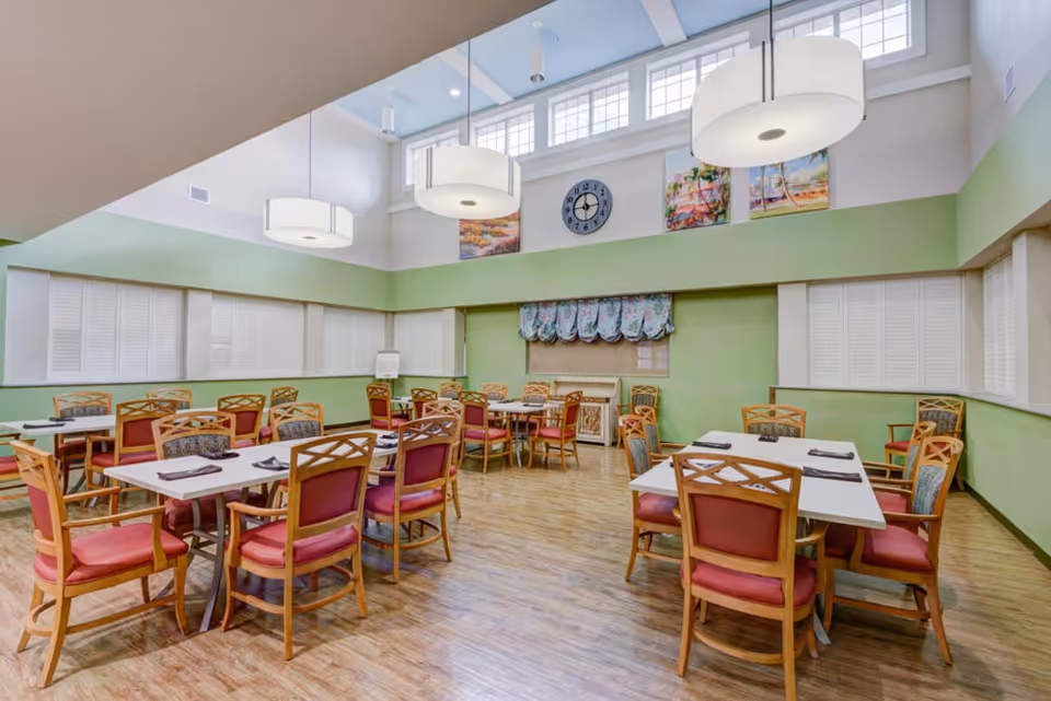 A bright dining room with multiple tables and wooden chairs with red cushions. The walls are painted light green with white trim, and there are large windows near the ceiling letting in natural light. Three large round pendant lights hang from the ceiling. A clock and two colorful paintings are mounted on the wall above a window with floral curtains.