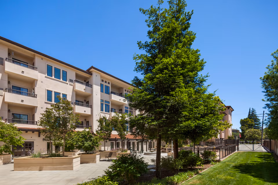 Outdoor courtyard area of a multi-story senior living facility with balconies, trees, shrubs, and a paved walkway under a clear blue sky.