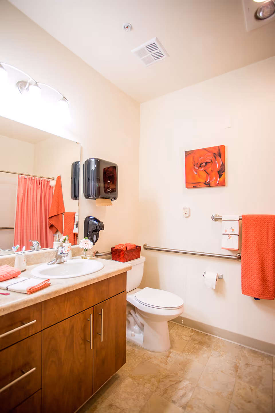 A clean and well-lit bathroom with a wooden vanity cabinet, a white sink, and a large mirror above it. There are red towels hanging on a grab bar next to the toilet, which has a red basket with rolled towels on top. A red shower curtain is visible in the reflection of the mirror. The walls are light-colored, and there is a red floral painting hanging above the grab bar. The floor is tiled in a beige pattern.
