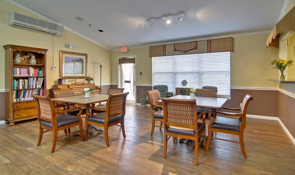A well-lit dining room with two wooden tables, each surrounded by four wooden chairs with cushioned seats. There is a wooden bookshelf filled with books and decorative items on the left side, and a piano beneath a framed picture on the wall. Large windows with blinds and a valance let in natural light. The room has wooden flooring and light yellow walls with a brown wainscoting. A door with a sheer curtain is visible in the background.