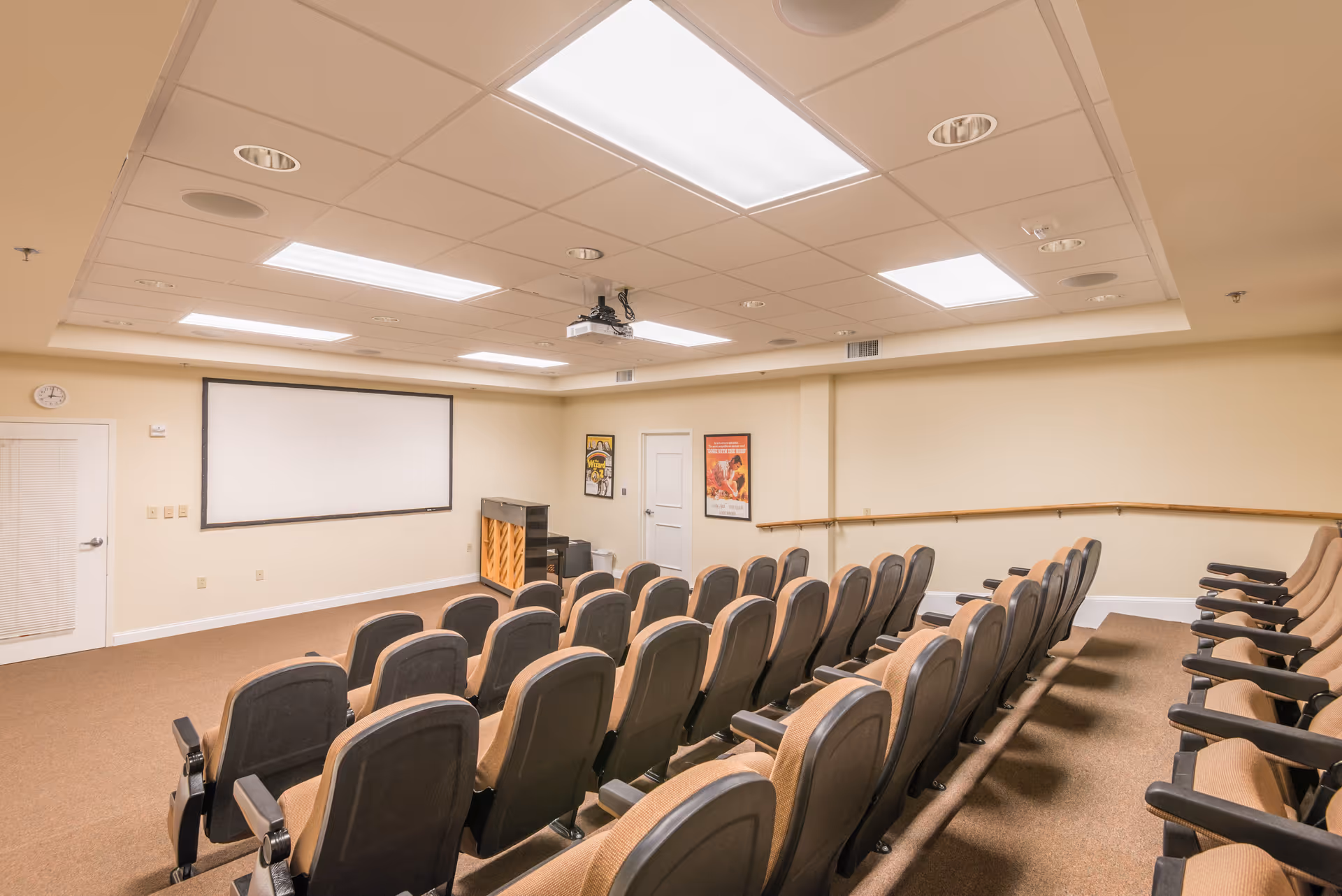 A small theater-style room with multiple rows of cushioned seats facing a large white projection screen on the wall. The room has beige walls, a carpeted floor, ceiling lights, a projector mounted on the ceiling, a piano, and framed posters on the walls.