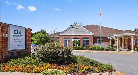 Exterior view of a senior living facility named Dimensions Living Prospect Heights, showing a brick building with a peaked roof, an American flag on a flagpole, a covered entrance, and a landscaped garden with colorful flowers in the foreground under a blue sky with some clouds.
