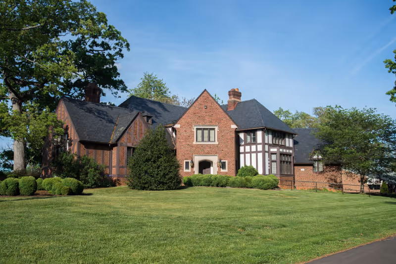 A large brick and timber Tudor-style building with multiple gables and chimneys, surrounded by green grass, bushes, and trees under a clear blue sky.