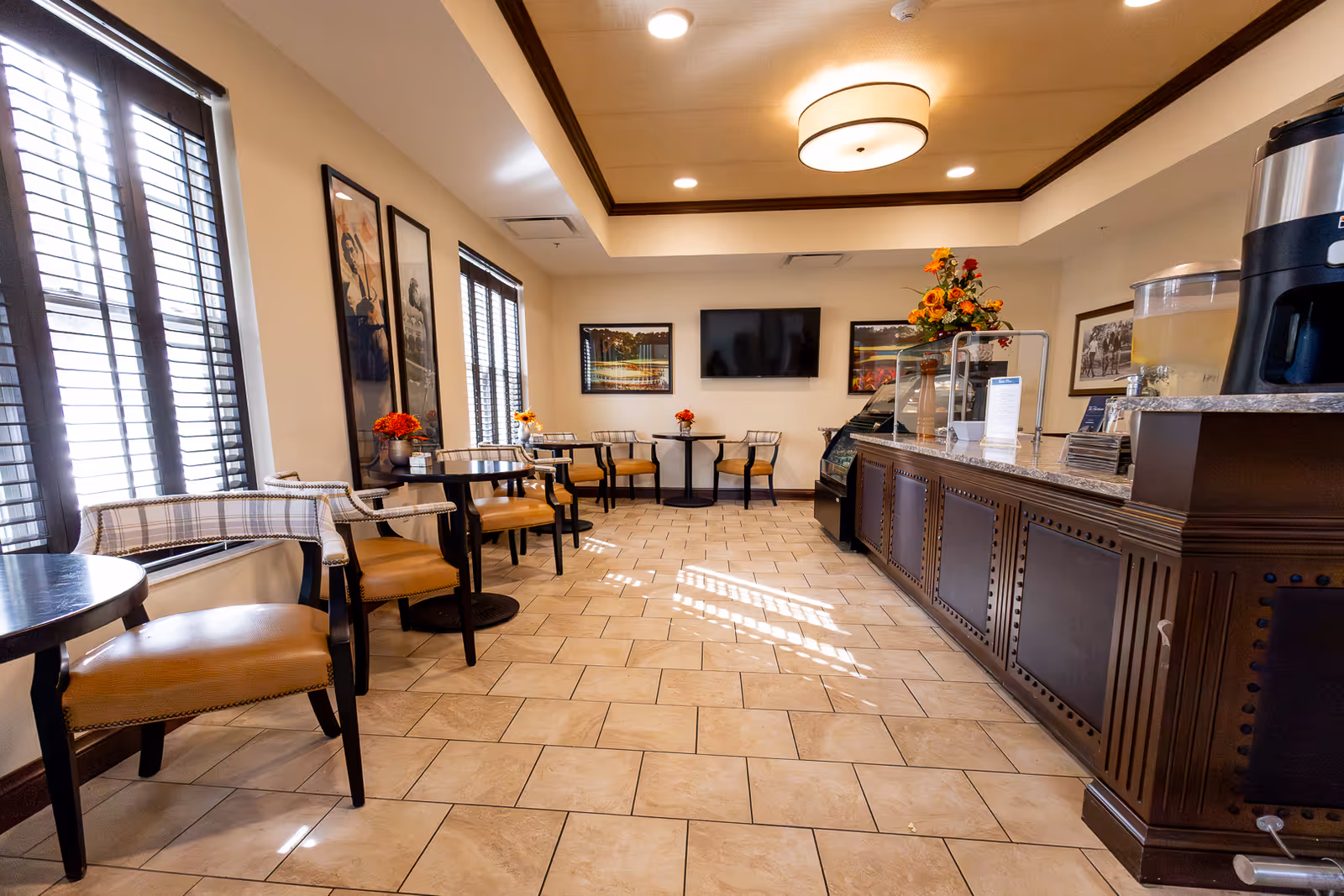 A bright dining area with several small round tables and chairs arranged along the windows and walls. The room features large windows with blinds, framed artwork on the walls, a flat-screen TV mounted on the far wall, and a counter with a beverage dispenser and flowers on top. The floor is tiled, and the ceiling has recessed lighting with a central circular light fixture.