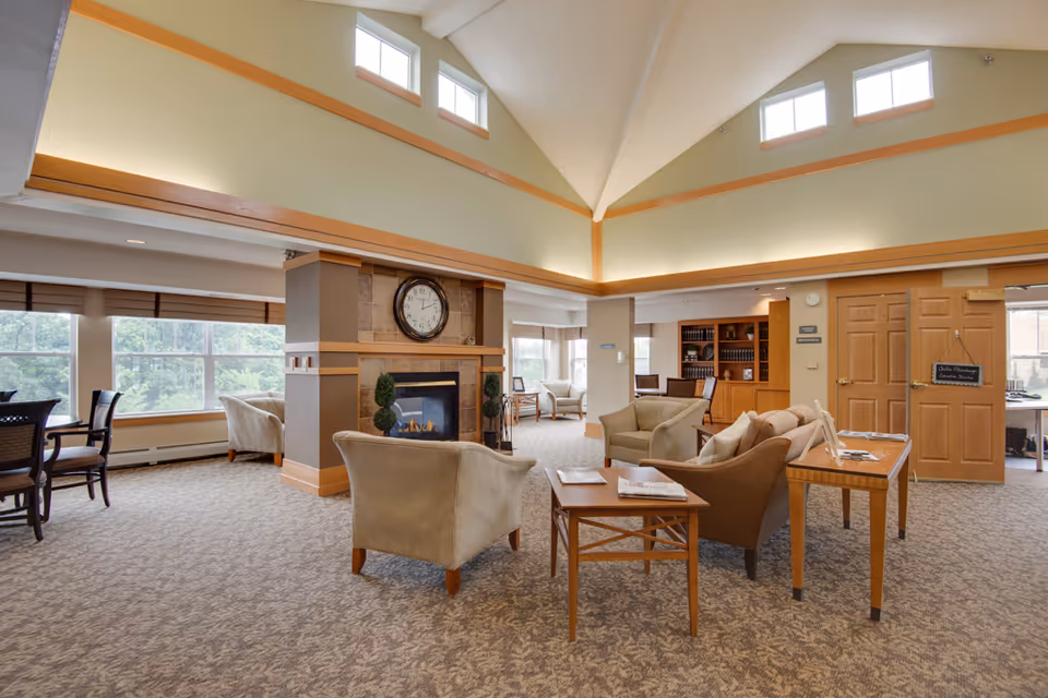 A spacious and well-lit living room area in a senior living facility with high vaulted ceilings and multiple small windows near the ceiling. The room features a central fireplace with a large clock above it, surrounded by comfortable armchairs and sofas arranged around coffee tables. Large windows on one side allow natural light to fill the space, and there is a wooden bookshelf and a table with informational materials near a set of double doors.