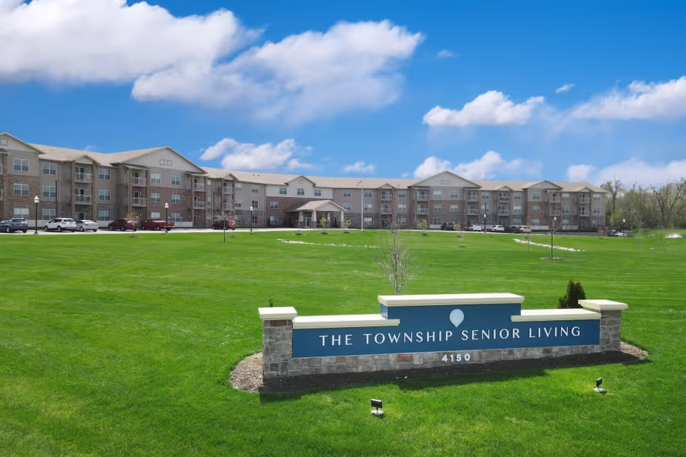 Well-kept green lawn and entrance sign in front of The Township Senior Living multi-story building under a blue sky.