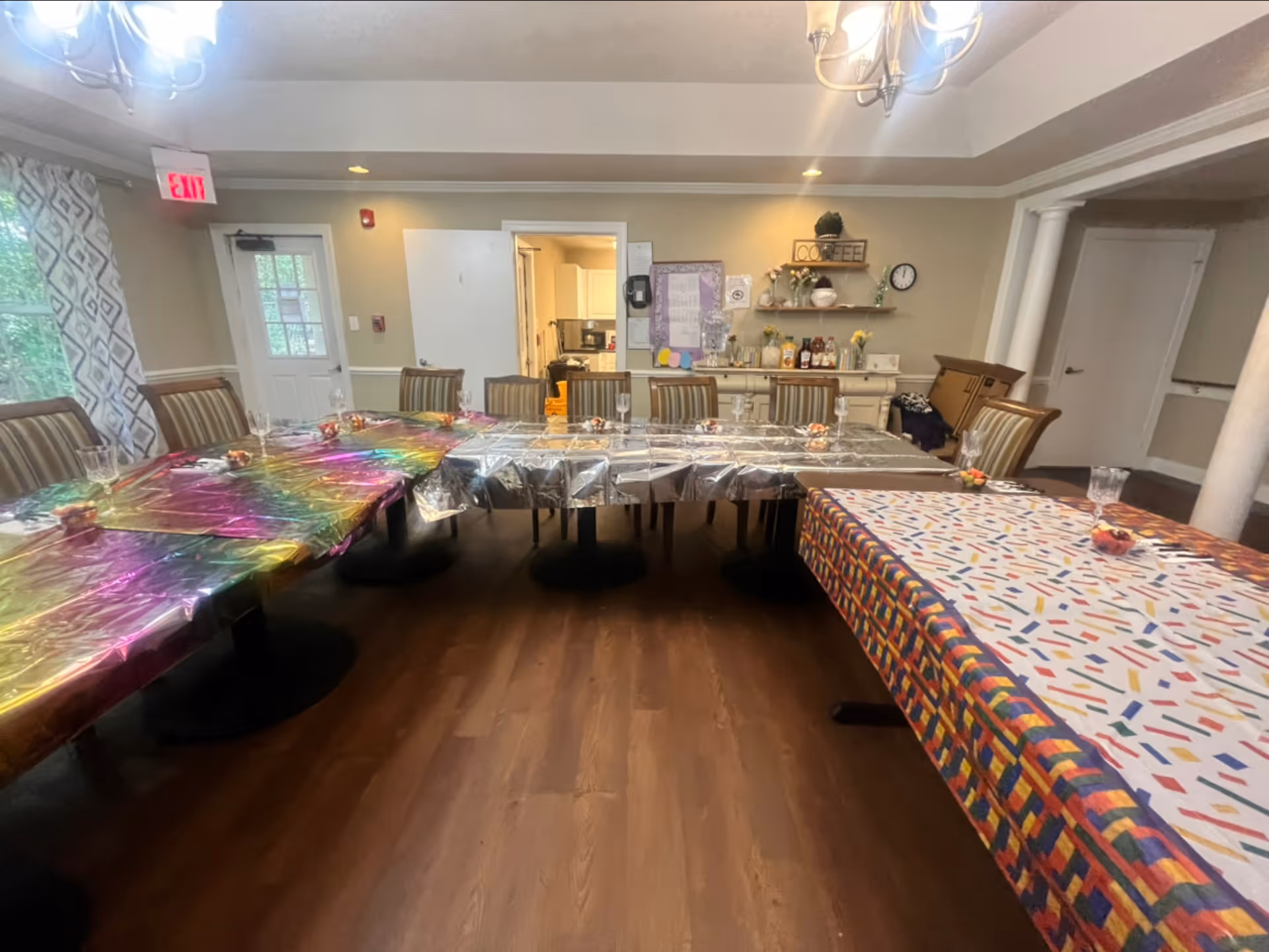 A communal dining room with long tables covered in colorful tablecloths, chairs around them, and a coffee station along the back wall.