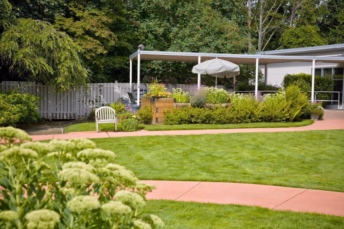 A well-maintained outdoor garden area with green grass, a curved pinkish pathway, various shrubs and flowering plants, a white wooden chair, and a covered patio with an umbrella and seating. Trees and a white fence are visible in the background.