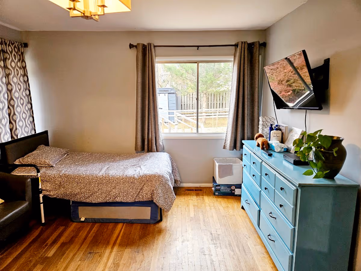 Bright bedroom with a single bed, blue dresser, wall-mounted TV and a window overlooking a fenced yard.