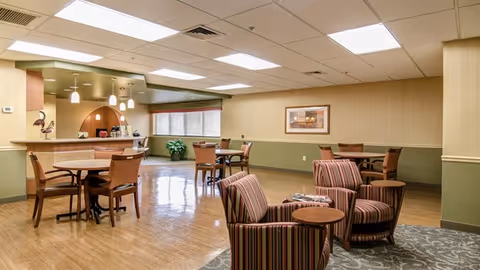 A spacious common area in a senior living facility featuring several wooden tables with chairs and a small seating area with striped armchairs and a round wooden table. The room has a polished wooden floor, beige walls with a green lower half, and a ceiling with recessed lighting. A framed picture hangs on the wall, and a counter with pendant lights is visible in the background.