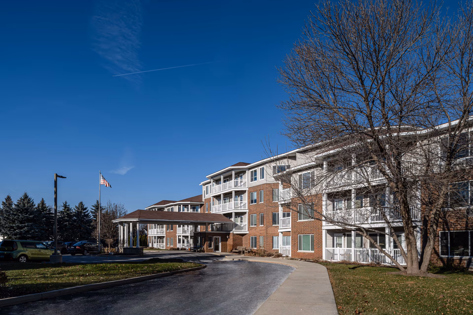 Exterior view of a multi-story senior living facility building with brick and white siding, balconies, a covered entrance, a driveway, and a flagpole with an American flag under a clear blue sky.
