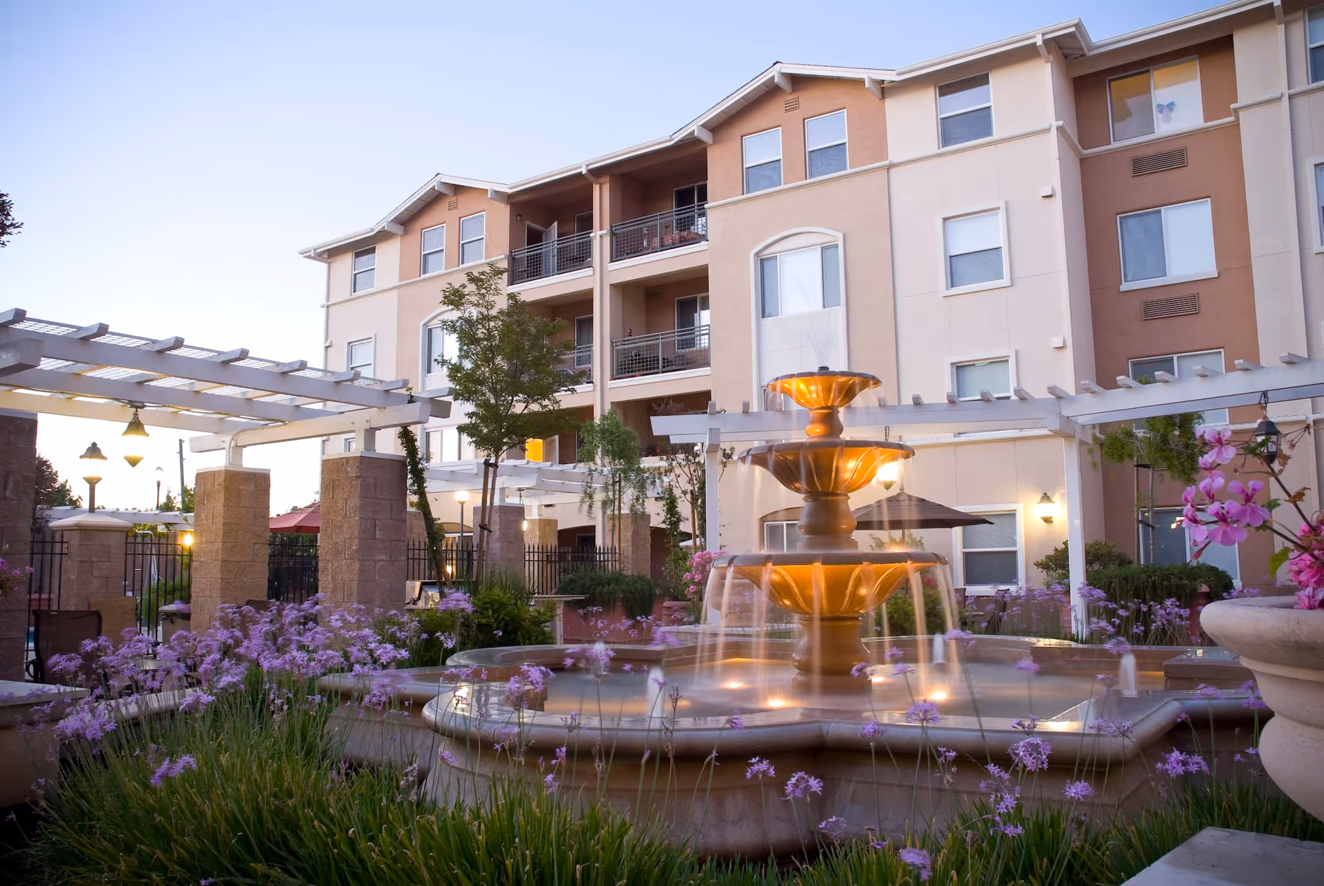 Outdoor courtyard area of Heritage Estate Senior Apartments featuring a three-tiered illuminated fountain surrounded by purple flowers and greenery, with a multi-story residential building in the background and white pergolas with stone pillars.