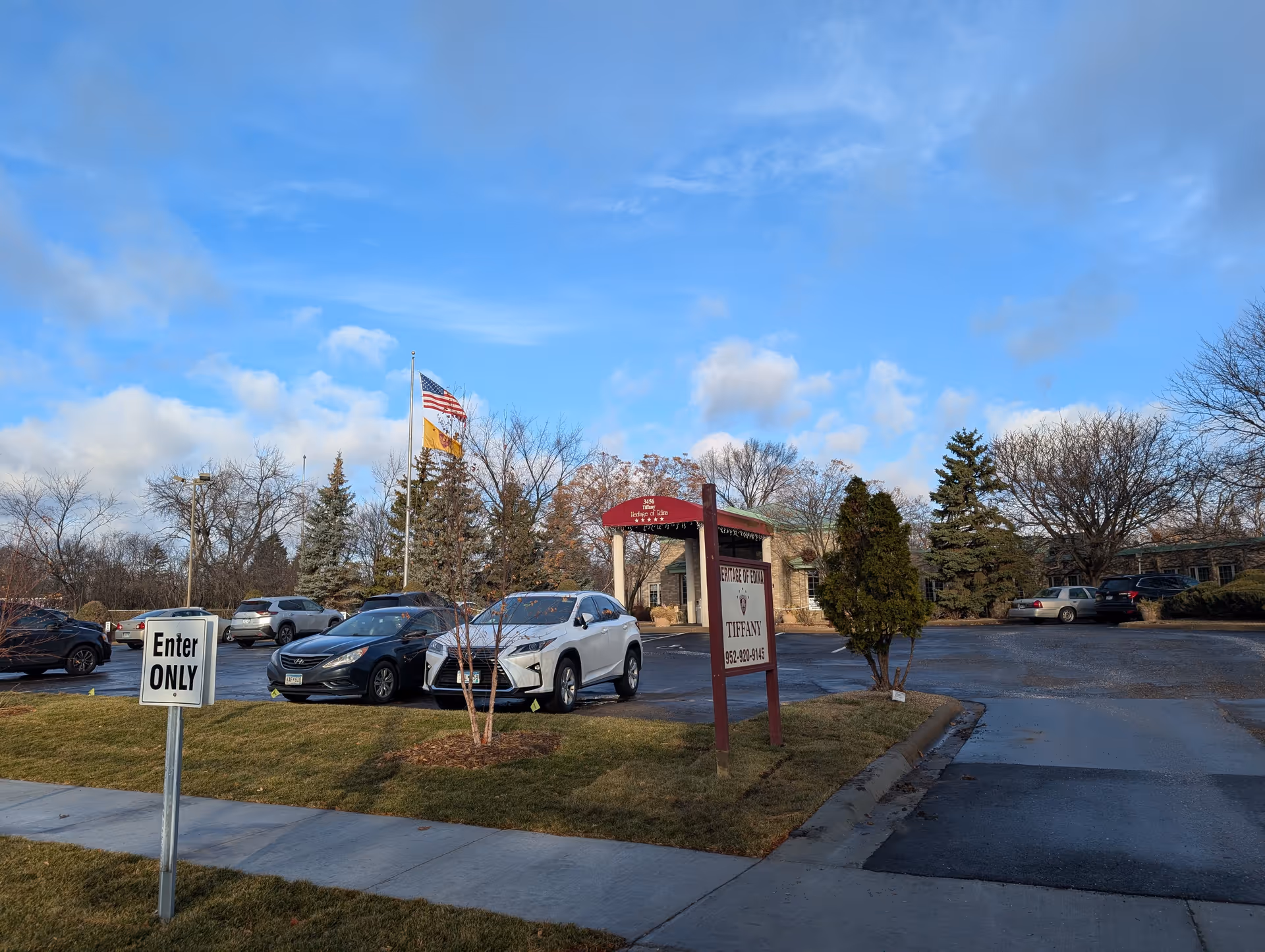 Exterior view of Heritage Of Edina II senior living facility parking lot with several parked cars, a sign displaying the facility name and contact number, two flagpoles with American and another flag, and a clear blue sky with some clouds.