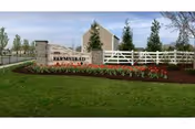 Stone entrance sign reading "The Farmstead" with a white fence, flowerbeds, and buildings behind it.