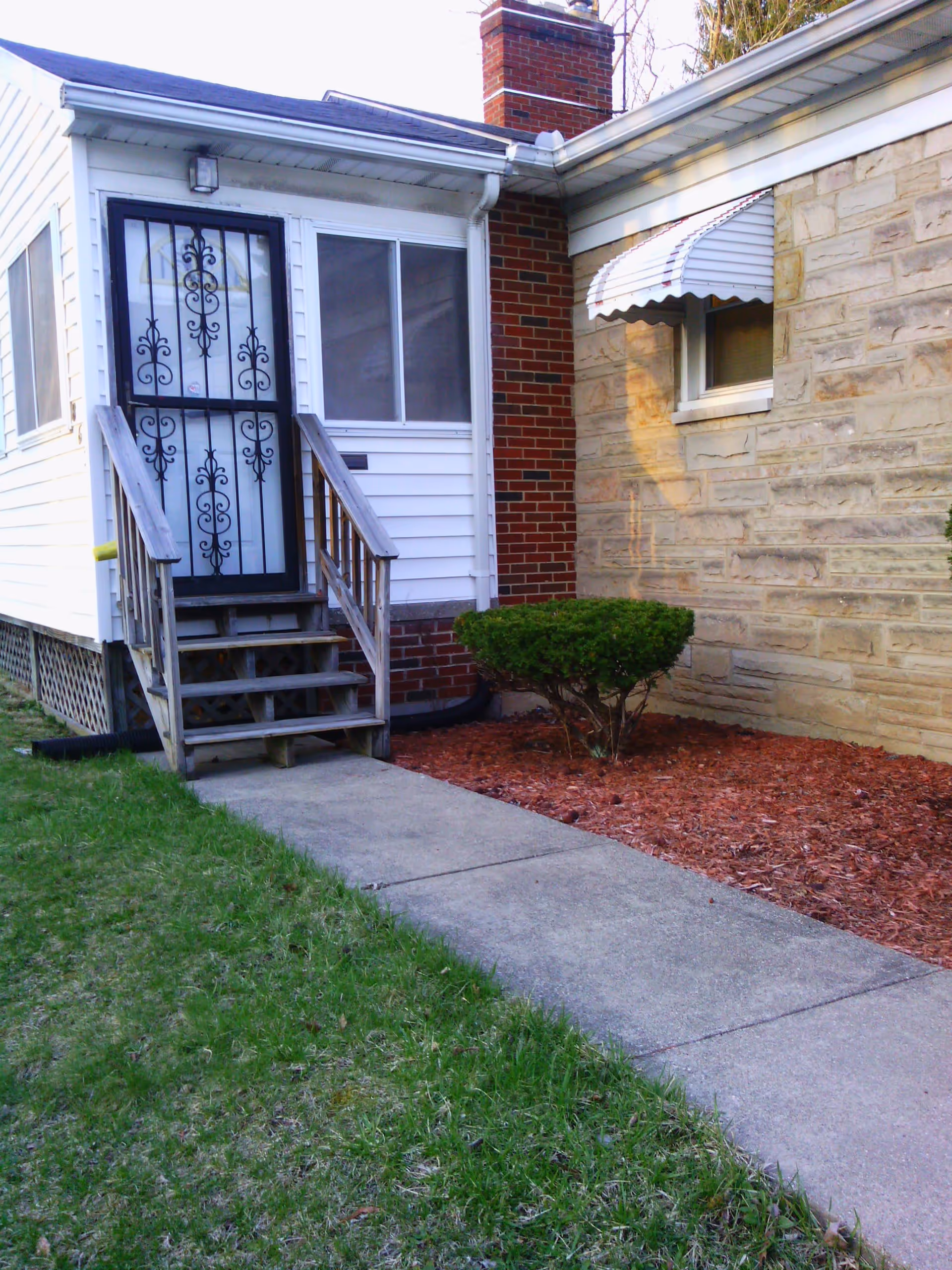 Exterior view of a house entrance with a small wooden staircase leading to a black decorative metal door. The house has white siding and a brick chimney, with a small bush and mulch bed next to a stone wall with a window covered by a white awning. A concrete walkway leads up to the stairs, bordered by grass on one side.